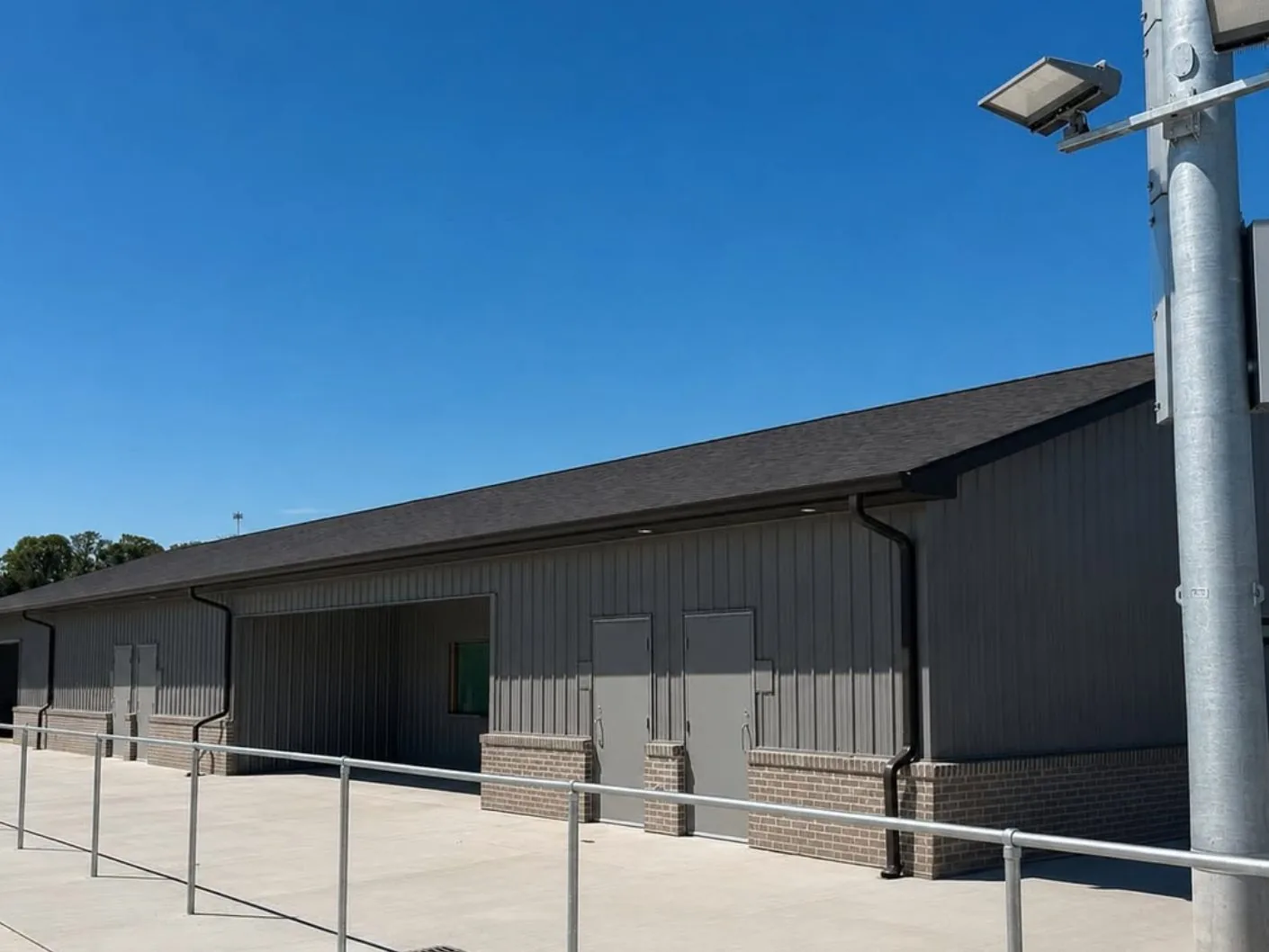 Long gray metal-sided building with brick wainscot, black gutters and downspouts and a shingle roof at a sports complex